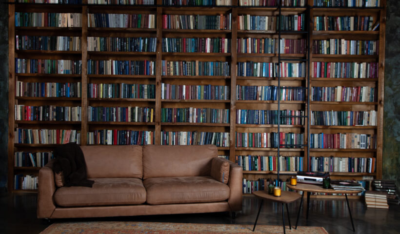 Dark Academia Fall Aesthetic Dark brown leather couch with blanket over it against a wall of dark brown bookshelves, next to small tables with candles and media players, Shutterstock, Real_Life_Photo