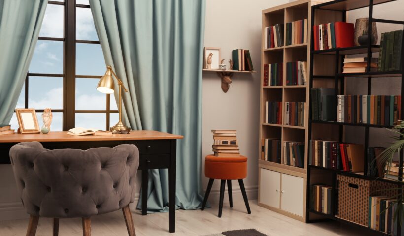 home office work environment with brown and black desk, plush tufted mauve chair, sage green curtains, modern bookshelves, orange stool with books on it, brass lamp