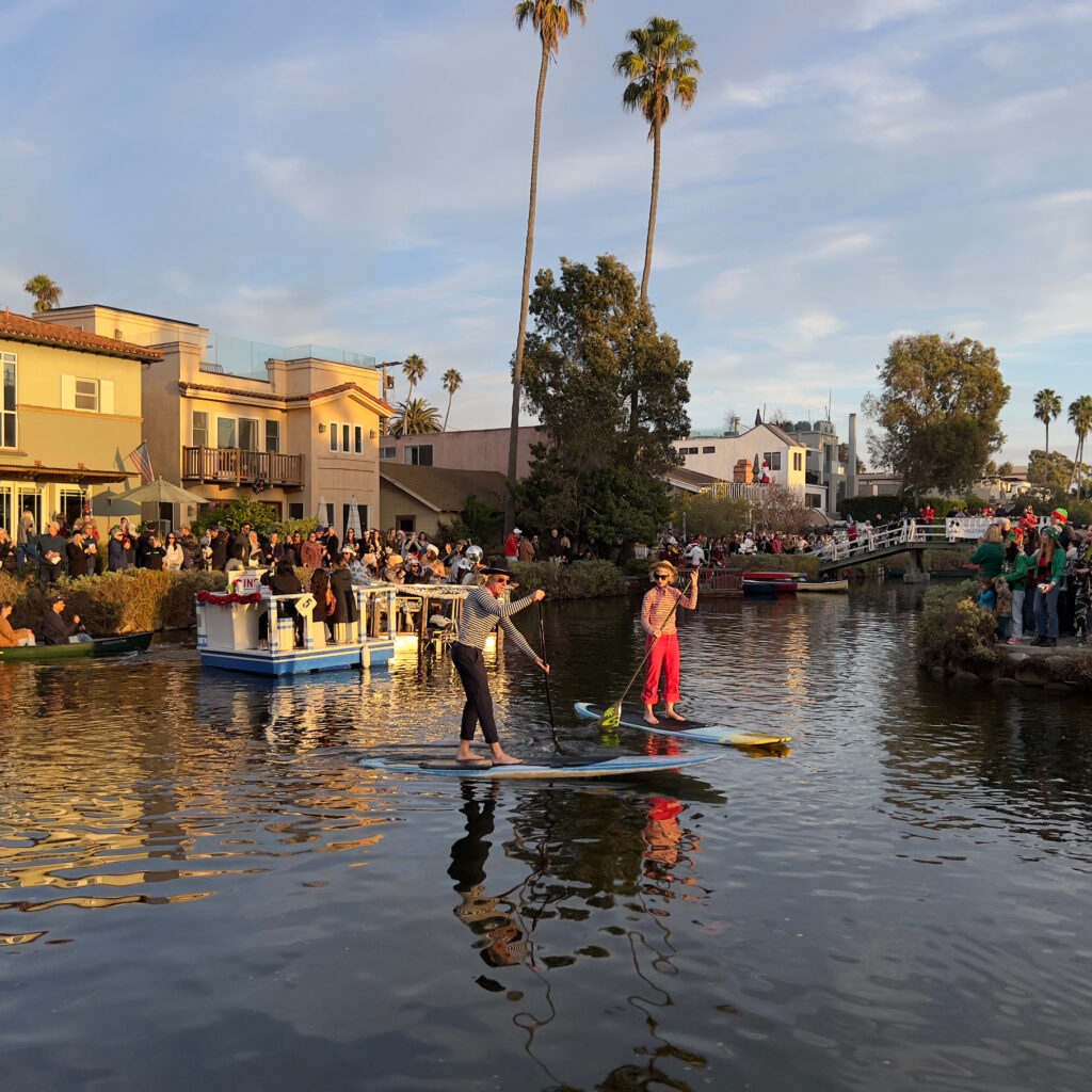 Gondoliers on paddle boards with palm trees and diner float in the background Venice Canal Boat Parade Christmas Decorations Holiday Decorations California Celebration